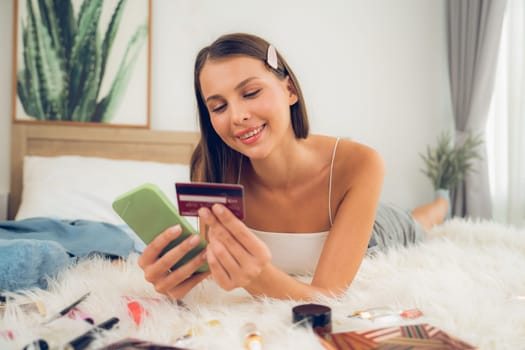 Young woman lying on the bed using online payment app and digital wallet on smartphone to pay with credit card. E commerce shopping and modern purchasing via mobile internet. Unveiling