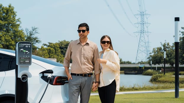 Young couple recharge EV car battery at charging station connected to power grid tower electrical industrial facility as electrical industry for eco friendly vehicle utilization. Expedient