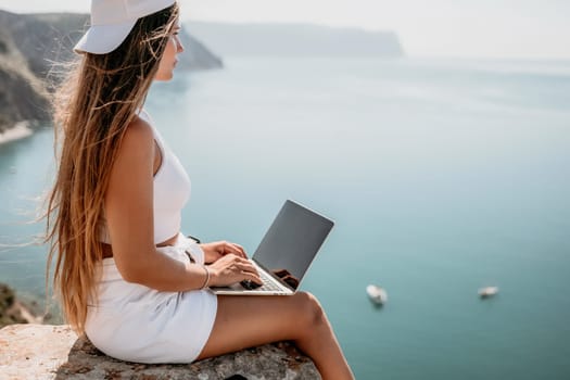 Successful business woman in yellow hat working on laptop by the sea. Pretty lady typing on computer at summer day outdoors. Freelance, travel and holidays concept.