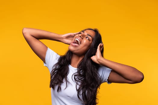 Happy young Indian woman wearing wireless headphones singing song out loud. Isolated on yellow background. Studio shot. Lifestyle concept.