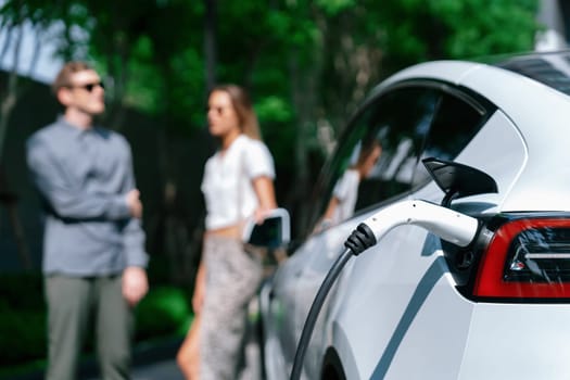 Young couple travel with EV electric car charging in green sustainable city outdoor garden in summer shows urban sustainability lifestyle by green clean rechargeable energy of electric vehicle innards