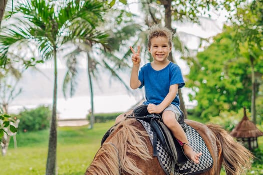 Blond boy riding horse in hotel park on the beach. Sunny summer day happy childhood.