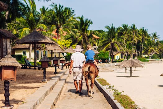 Blond boy riding horse in hotel park on the beach. Sunny summer day happy childhood.