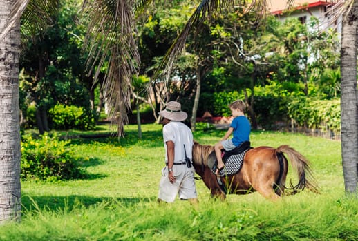 Blond boy riding horse in hotel park on the beach. Sunny summer day happy childhood.