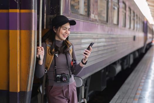 Cute Asian traveler playing with phone, carrying backpack, waiting for train at train station.