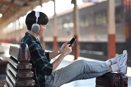 Young man in headphones using mobile phone while sitting on bench at railway station.