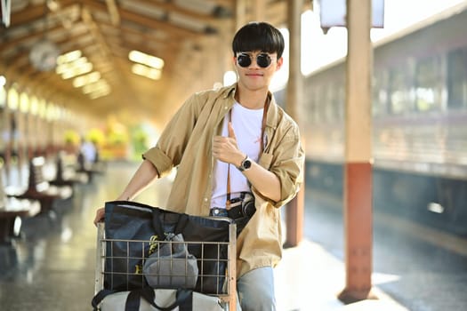 Young man wearing sunglasses walking with luggage cart on the corridor at railway station.
