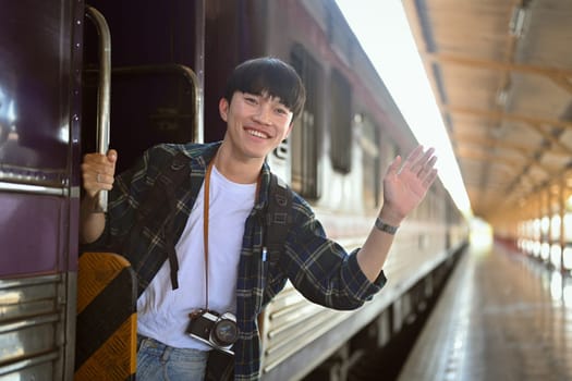 Happy young Asian man waving hand from the train while leaving the station. Traveling concept.