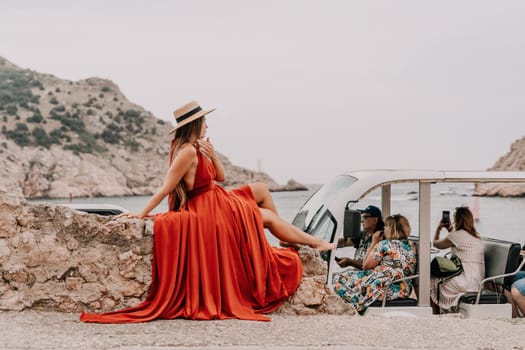 Side view a Young beautiful sensual woman in a red long dress posing on a volcanic rock high above the sea during sunset. Girl on the nature on overcast sky background. Fashion photo