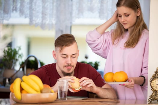 A man is sitting at the kitchen table eating a sandwich. In the background you can see a window with a curtain, a kitchen countertop, a sink and various kitchen appliances. A bowl of fruit stands next to the man.