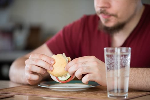 A man sits at a table. In front of him stands a plate and a glass of water. He holds a vegetable sandwich in his hands. Blurred background.