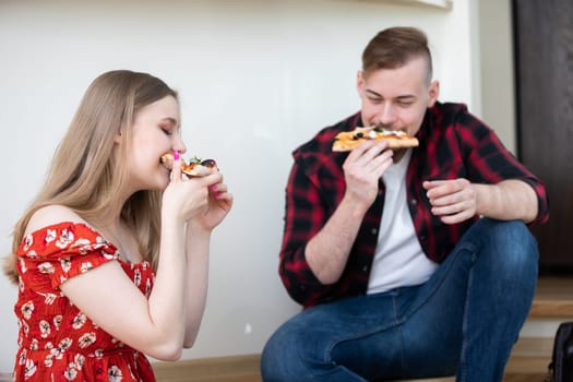 A quick meal eaten on the run. Friends take a break and greedily eat pizza. They sit on the floor. Each of them is holding a piece of pizza.