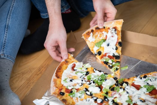 The boy reaches for two pieces of pizza. Only his hands are visible. Two pairs of legs can be seen in the background. Two people are sitting on the floor eating pizza.