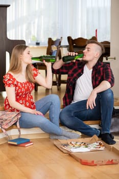 A man and a woman sit on the dining room floor. A pizza box lies in front of them. They are facing each other and each is drinking from a glass green bottle.