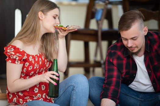 A hungry young girl bites into a slice of pizza with her eyes closed. She holds a bottle of beer in her other hand. Next to her sits a man with a beard and mustache.
