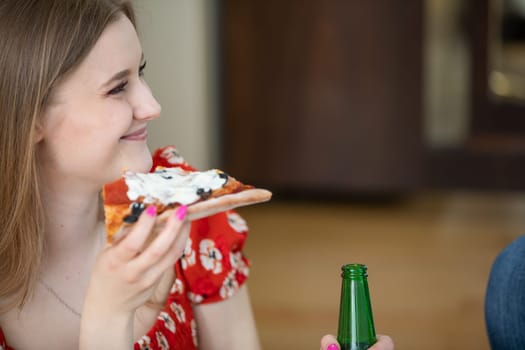 The figure of a young woman seen in profile. She is smiling and holding a slice of pizza. In her other hand the woman is holding a glass bottle of beer.