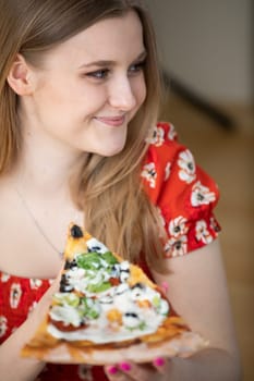 The girl is wearing a red floral shirt. She wears a silver necklace around her neck. A smiling young blonde woman holding a slice of pizza.