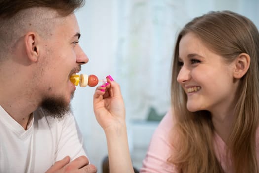 The girl feeds the man fruit. A grape, a slice of banana and an orange are impaled on a toothpick. The boy eats from his girlfriend's hand.