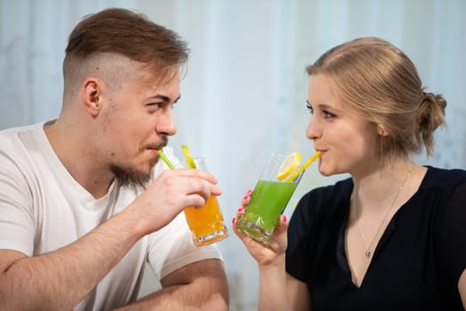 A couple in love looks into each other's eyes. They drink colorful drinks with straws. The woman has an elegant hairstyle and a black dress. The man is dressed in a white T-shirt.