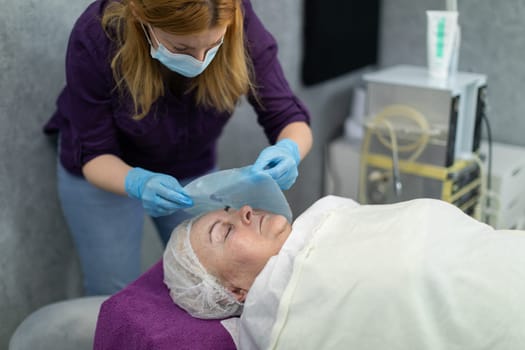A beautician works providing services in a beauty salon. She places a cloth soaked in a moisturizing substance on a client's face. In the background you can see gray walls and shelves of cosmetics.
