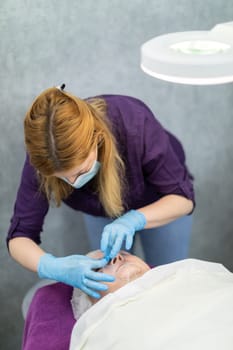 A young woman works in a beauty salon as a cosmetologist. She attends to her client, who is lying next to her on the cosmetic bed.