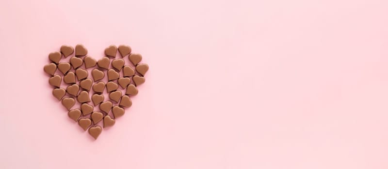 Heart made of romantic chocolate confections in heart shapes on pink background. Long horizontal banner with chocolate heart of milk chocolate sweet, copyspace. Topview flatlay. Valentines Day concept