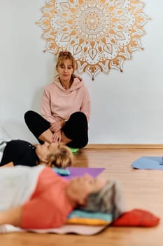 A skilled trainer oversees a group of senior women practicing various yoga exercises, including neck, back, and leg stretches, in a sunlit space, promoting wellness and harmony.