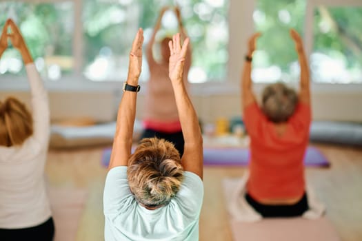 A group of senior women engage in various yoga exercises, including neck, back, and leg stretches, under the guidance of a trainer in a sunlit space, promoting well-being and harmony.