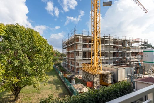 Construction site under construction. In the background blue sky and white clouds. Around large garden with large tree.