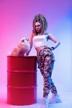 cool girl with braided dreadlocks on her head with a cute Pomeranian dog in neon light on a light background