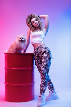 cool girl with braided dreadlocks on her head with a cute Pomeranian dog in neon light on a light background