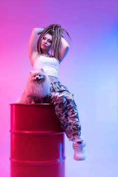 cool girl with braided dreadlocks on her head with a cute Pomeranian dog in neon light on a light background