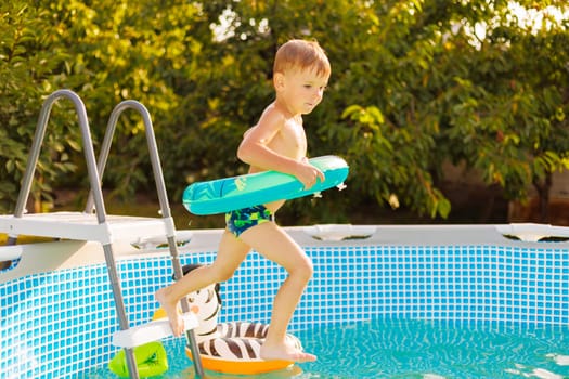 A little boy is ready for pool time fun, stepping into the cool water with his bright inflatable toy on a sunny day surrounded by lush greenery.