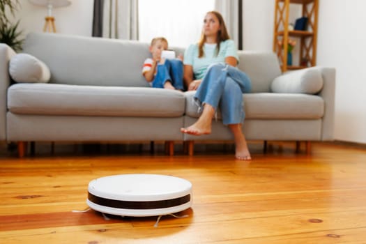 A robotic vacuum cleaner cleans the floor as a woman relaxes with a child on the sofa in the background
