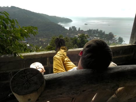 a man sits on a wooden chair admiring the view of the sea and mountains from a height.