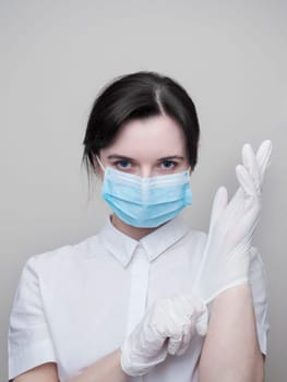 Young woman patient in a medical mask puts on protective surgical sterile gloves on her arm, on gray background, protection against coronovirus