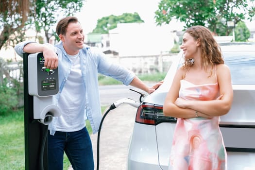 Happy and lovely couple with eco-friendly conscious recharging electric vehicle from EV charging station. EV car technology utilized as alternative transportation for future sustainability. Synchronos