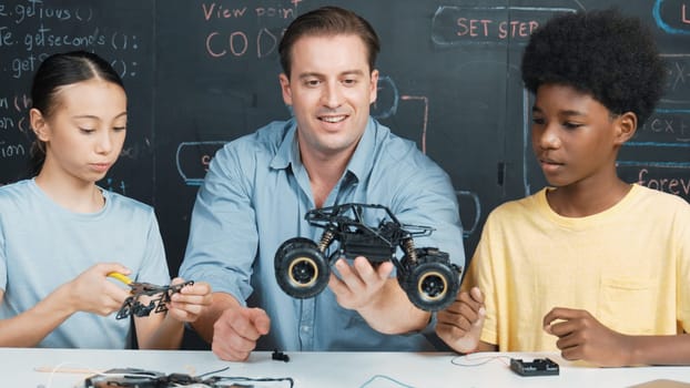 Smart teacher inspect car model construction to diverse student. Highschool children with mixed races listening mentor explain robotic model system at table with laptop and wires placed. Edification.