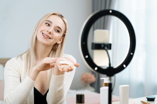 Young woman making beauty and cosmetic tutorial video content for social media. Beauty blogger smiles to camera while showing how to apply beauty skincare to audience or followers. Blithe