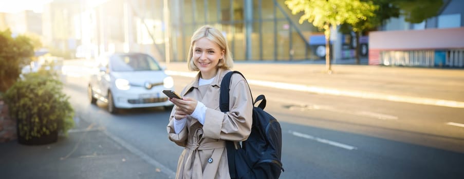 Stylish young blonde woman, girl with smartphone and backpack, standing on street, using mobile phone application, posing close to the road with cars and sky scrappers.