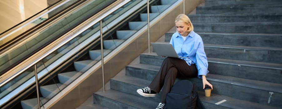 Freelance employee, woman working on her project, using laptop, sitting outdoors on stairs, drinking takeaway coffee.