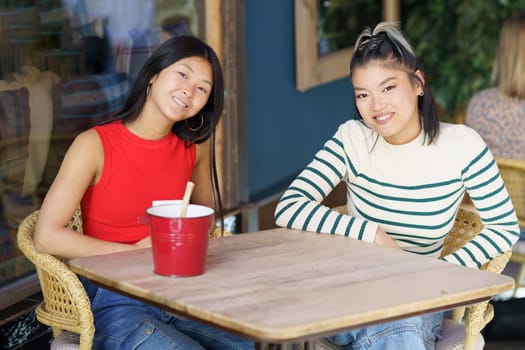 Smiling young Asian girlfriends in casual clothing sitting together at wooden table and discussing plans while spending time in cafe