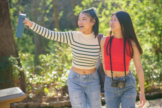 Cheerful young Asian female friends in casual apparel, standing in green forest of Alhambra and pointing away while enjoying picturesque views during summer trip in Granada