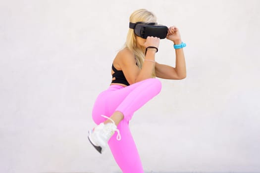 Young blond haired female athlete in sportswear and VR goggles practicing punches and kicks during virtual boxing training against white background