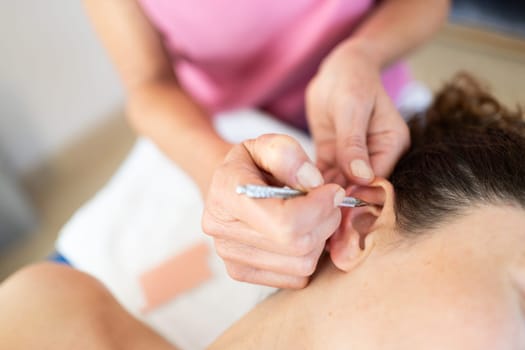 Crop professional beautician applying auriculotherapy ear acupuncture techniques of female client lying in modern beauty salon during facial treatment