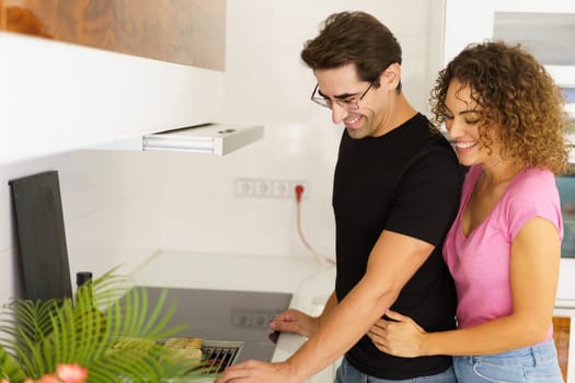 Side view of happy adult couple looking down while standing in kitchen near cooking range with bread slices, on grill and female embracing male in eyeglasses from behind in daylight