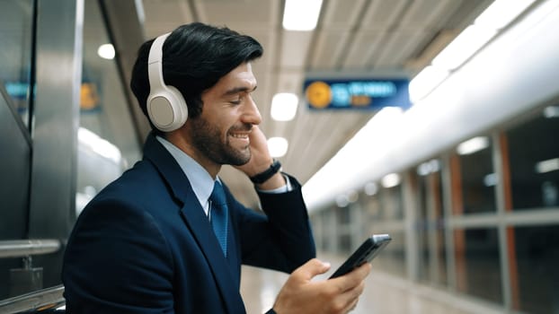 Smart business man listening music by headphone while waiting train at train station with blurring background. Skilled project manager enjoy listen relax sound while holding mobile phone. Exultant.