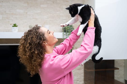 Side view of happy young female in pink casual cloth with curly hair lifting adorable black and white cat, while playing and bonding in room at home