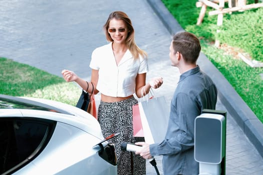 Young couple travel with EV electric car charging in green sustainable city outdoor garden in summer shows urban sustainability lifestyle by green clean rechargeable energy of electric vehicle innards