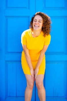 Delighted young female in yellow dress with curly brown hair bending forward and smiling while laughing with closed eyes against blue building on street